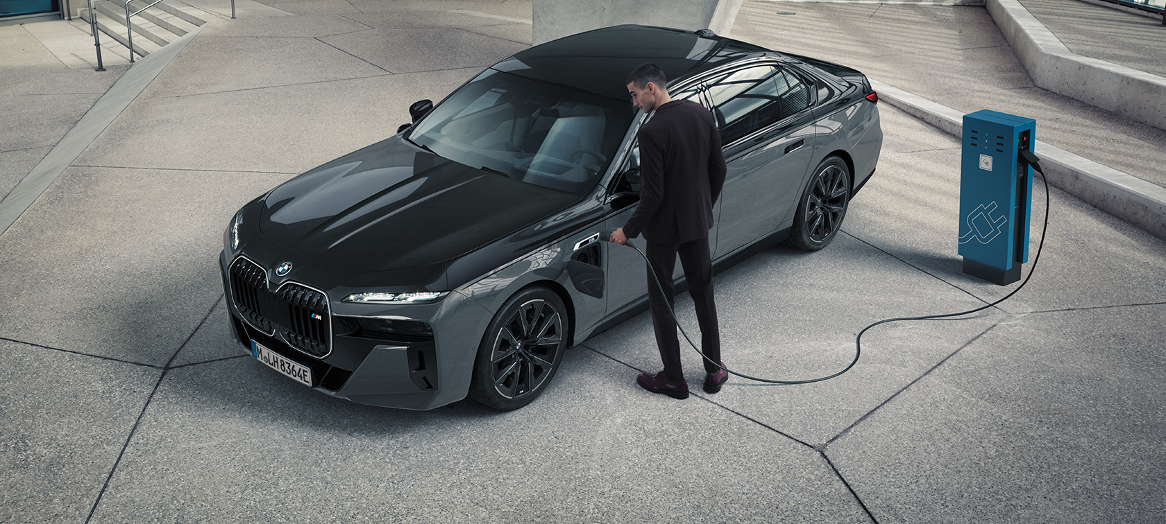 BMW M760e xDrive Sedan G70 M Performance package BMW M760e xDrive Sedan G70 M Performance Package BMW Individual two-tone Black Sapphire metallic paintwork BMW Individual Dravite Grey metallic three-quarter front view man with charging cable next to charging station