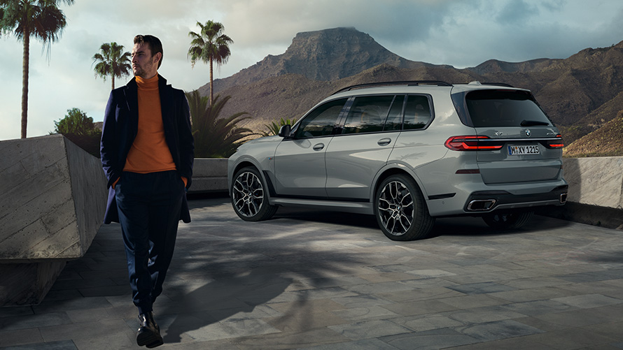 Man in front of parked BMW X7 (G07) Remote Control Parking Remote Control Parking man in front of parked BMW X7 (G07) three-quarter rear view beneath palm trees with mountain landscape as background
