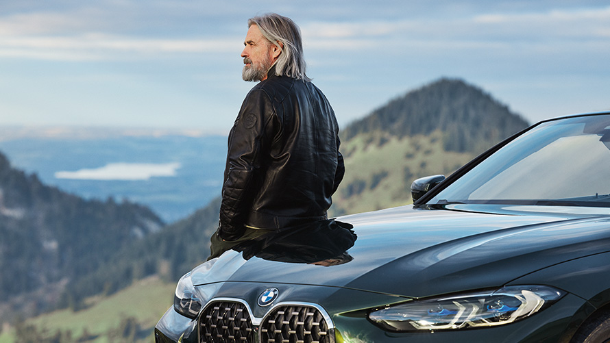 Man leaning against a BMW in front of mountain landscape BMW Spring Summer Accessories Man leaning against a BMW in front of mountain landscape
